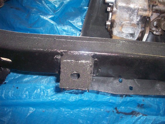 Black painted vehicle frame with a metal bracket and part of a drivetrain visible, resting on a blue tarp