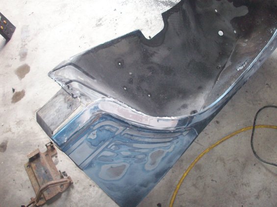 Blue vehicle fender section with body filler and primer, resting on a workbench