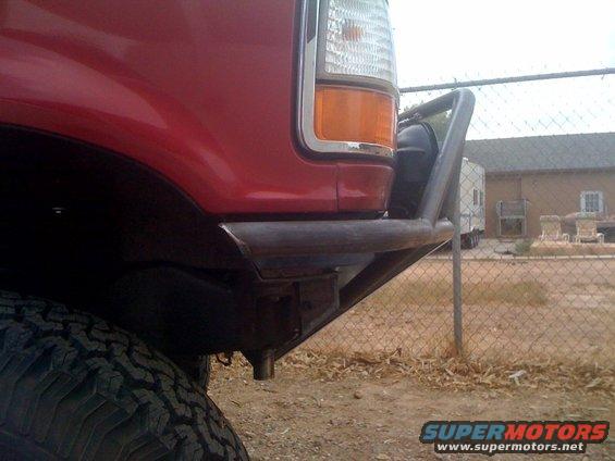 Red truck fender with aftermarket tubular bumper and off-road tire. Building and fence in background