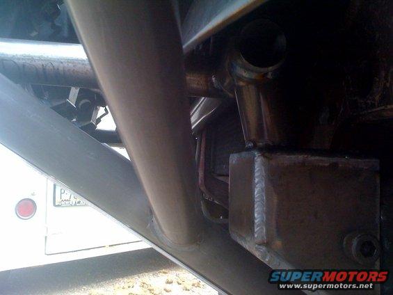 Close-up of vehicle frame and suspension parts, with a partial view of a white vehicle's rear and a license plate