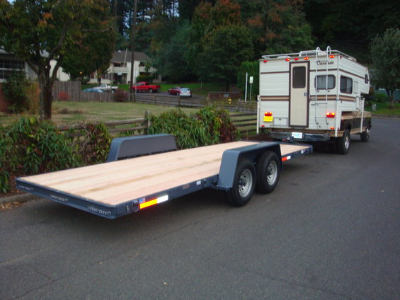 Flatbed trailer towed by a brown pickup truck with a white camper shell. The truck appears to be from the 1980s
