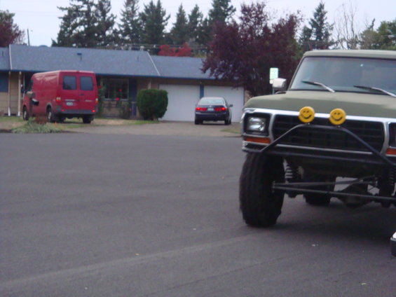 Green pickup truck with custom bumper and two yellow smiley face lights on the front. A red van and a dark sedan are parked in the background