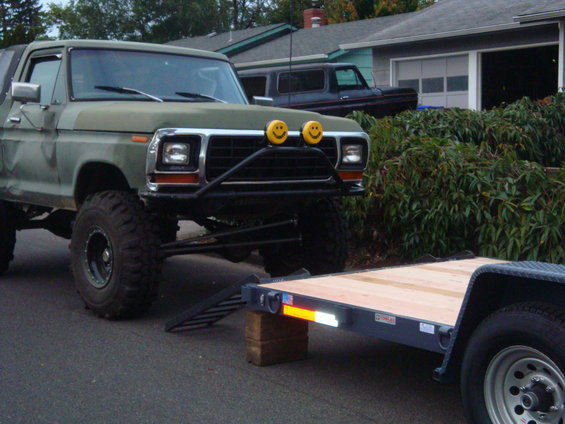 Lifted 1978 Ford Bronco with custom bumper and smiley face lights, parked next to a trailer. Another SUV is visible in the background