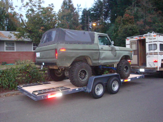 Olive green Ford Bronco with a black soft top, lifted suspension, and large off-road tires, loaded onto a dual-axle trailer