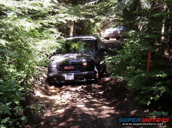 Black GMC SUV with red GMC logo on grille, driving on a muddy trail in a forest. Another vehicle is visible behind it