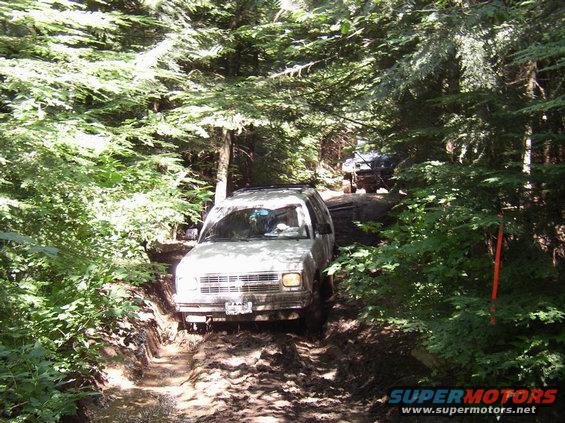Two SUVs driving on a muddy, wooded trail. The lead vehicle is light-colored and appears to be struggling on the terrain
