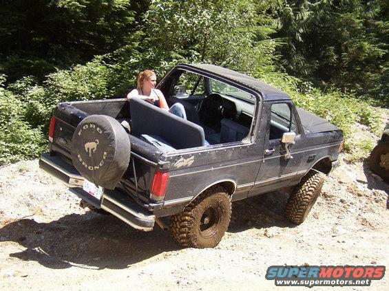 Black Ford Bronco with spare tire on back, driving off-road. Woman sits in the open rear
