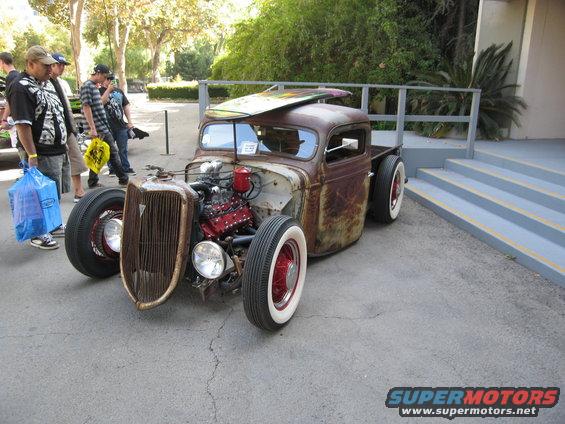 Rusty rat rod pickup truck with exposed engine and surfboard on roof