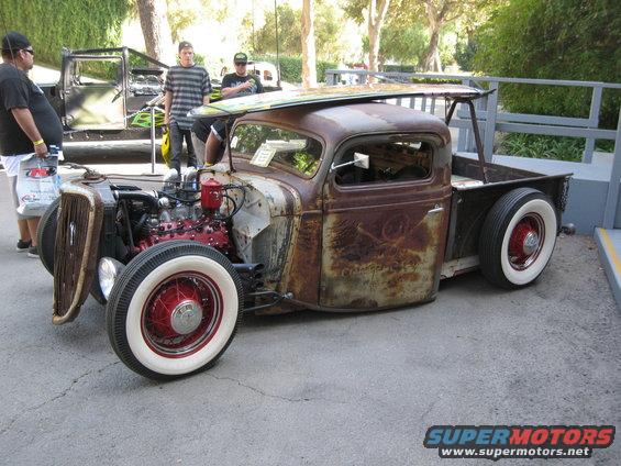 Rat rod pickup truck with a surfboard on top, red engine, white wall tires, and rusty body. Another truck is behind it