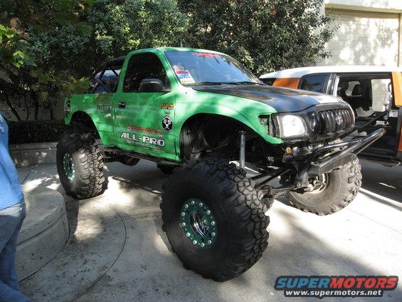 Green Toyota Tacoma pickup with large off-road tires and custom bumper. Another vehicle is partially visible behind it