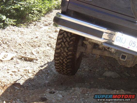 Dark SUV rear bumper with chrome trim and a muddy tire on a dirt road