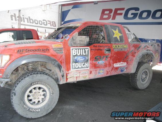 Red off-road racing truck with BFGoodrich tires and "Built Ford Tough" decal. Another red truck is partially visible behind it