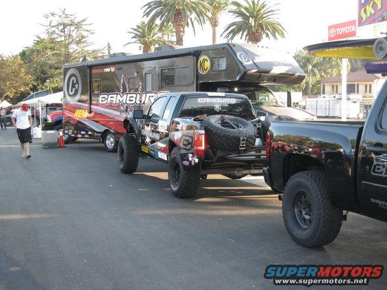 Modified pickup truck with spare tire mounted on rear, towing a large RV. Another pickup truck is parked to the right