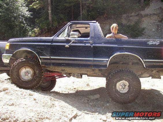 1988 Ford Bronco with lifted suspension and large tires on a dirt trail. A woman sits in the open rear