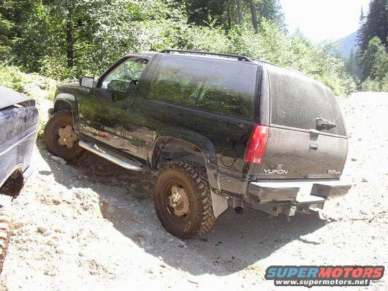 Black GMC Yukon SUV on a dirt road, rear 3/4 view. Off-road tires, side steps, and a roof rack are visible