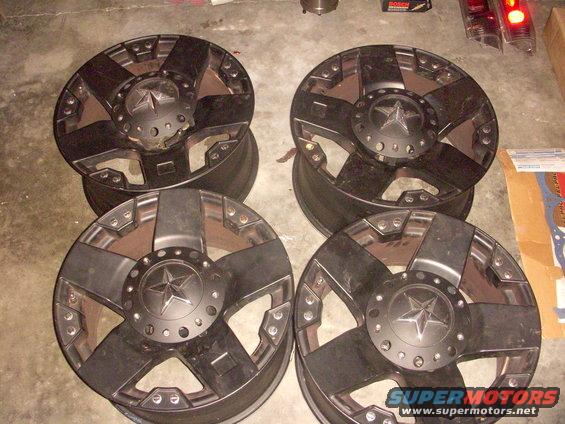 Four black aftermarket wheels with a star center cap stacked on a concrete floor
