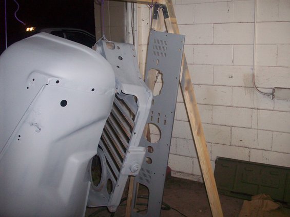 Gray Jeep CJ-5 grille and fender assembly hanging from a rack, with a metal panel leaning against it