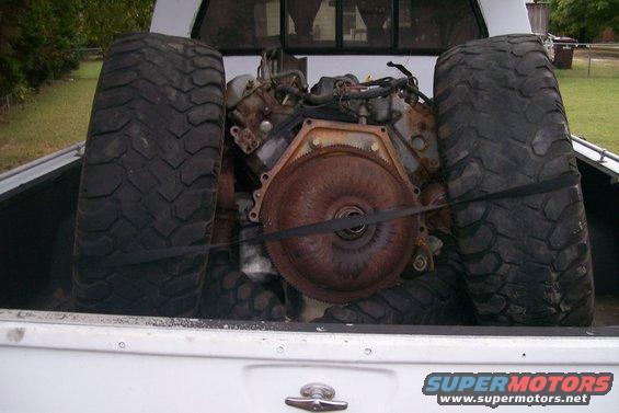 Engine and two large tires secured with straps in the bed of a white pickup truck