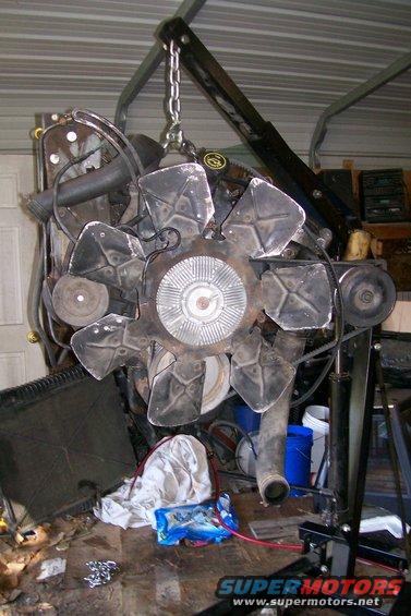 Engine assembly suspended by a chain hoist, with a large fan and belts visible