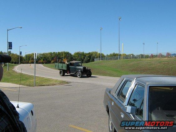 Vintage dark green flatbed truck driving on a road, with a modern car in the foreground