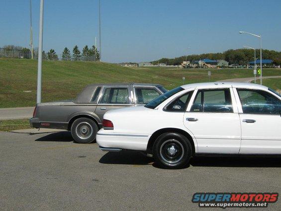 White sedan and a gray sedan parked on asphalt, with a grassy hill and buildings in the background