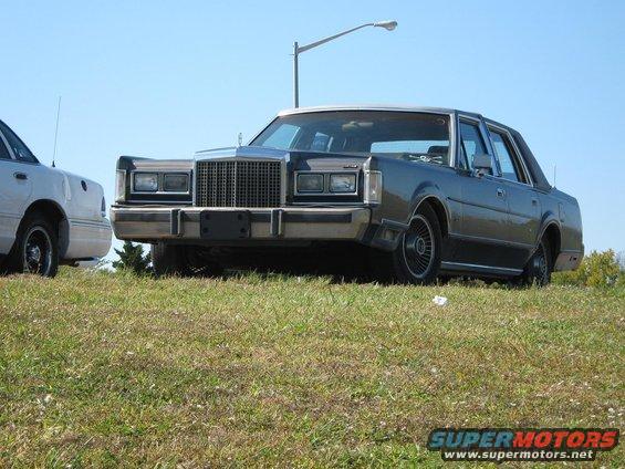 Dark gray Lincoln Town Car, front view, parked on grassy incline next to a white car