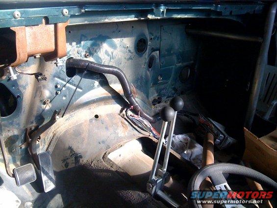 Interior of a vehicle with dashboard removed, showing exposed wiring, pedals, and a roll cage