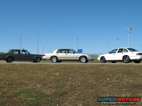 Three sedans parked on a grassy shoulder under a clear blue sky. The middle car is a tan 2000 Mercury Grand Marquis