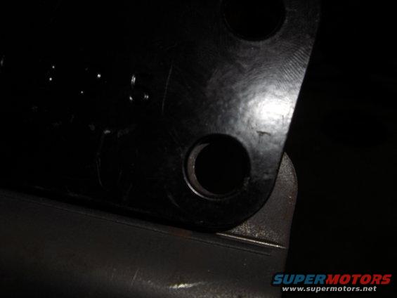 Black metal bracket with two circular holes, one partially visible. A SuperMotors logo is in the lower right corner