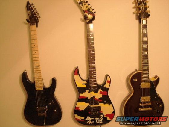 Three electric guitars hanging on a wall: a black one, a camouflage one with Japanese characters, and a dark brown one