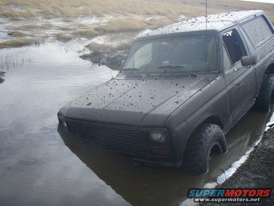 Dark gray Ford Bronco partially submerged in muddy water, front grille and tires visible