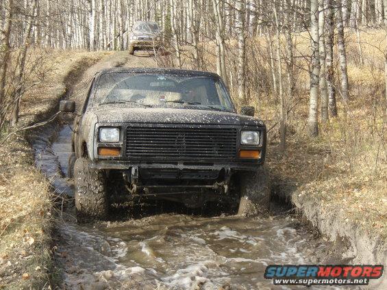 Muddy truck driving through a water-filled rut in a wooded area. Another vehicle is visible on the hill behind it