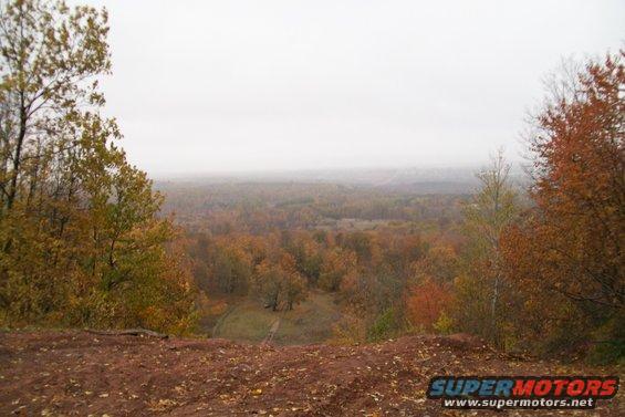 Overlook of a wooded valley in autumn with colorful trees and a hazy sky. Foreground is reddish dirt and fallen leaves