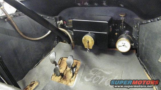 Interior view of a vintage car showing pedals, steering column, dashboard with gauges, and a floor mat with 'Ford' logo