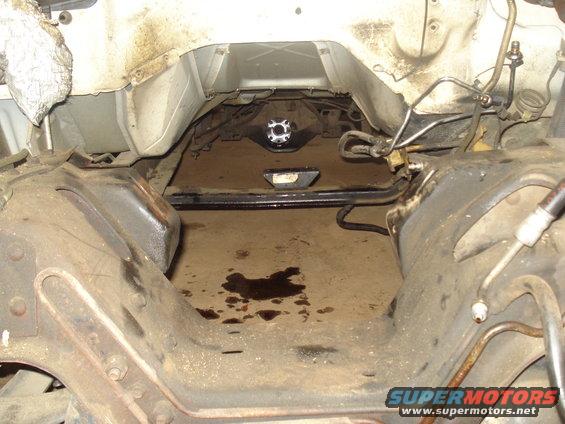 Underside of a vehicle showing the frame, transmission tunnel, and some drivetrain parts. There are oil stains on the floor