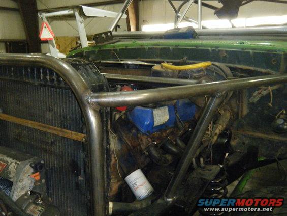 Green vehicle engine bay with custom roll cage, visible radiator, oil filter, and engine components