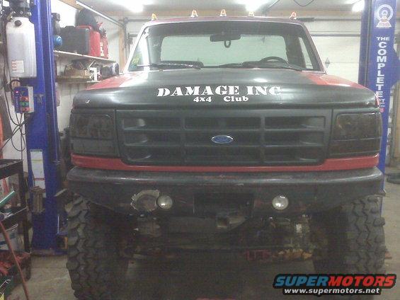 Front view of a red and black Ford F-Series truck with "DAMAGE INC 4x4 Club" on the hood, parked in a garage