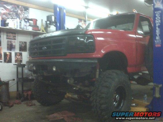 Red Ford truck with black hood and grille, lifted on large tires, in a garage with a lift