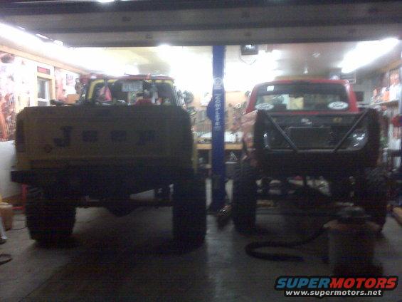 Two pickup trucks, one yellow with 'JEEP' on the tailgate and one red with a roll cage, parked in a garage