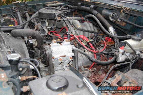 Engine bay with various hoses, wires, and engine components visible. Some rust is present