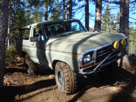 Olive green 1978 Ford Bronco with off-road tires and yellow smiley face headlights on a dirt trail
