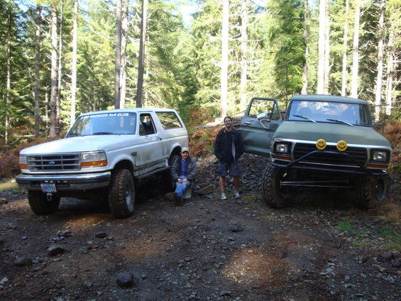Two people with a white Bronco and a green truck in a wooded area