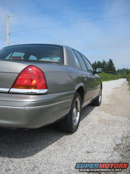 Rear quarter view of a grey sedan parked on a gravel road with trees and a blue sky in the background