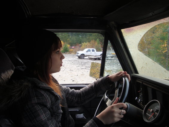 Woman with beanie and plaid jacket driving a 1978 Ford Bronco, looking out the window at a white pickup truck and trailer