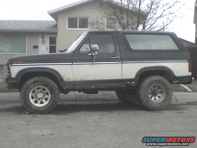 Side view of a two-tone Ford Bronco with a black top and white body. It has large off-road tires and is parked in front of a house