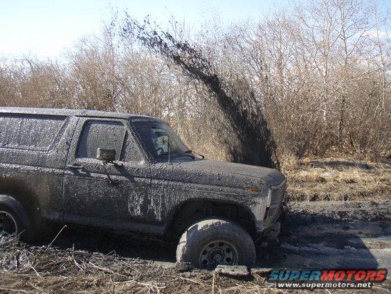 Mud-covered Ford Bronco stuck in mud, front quarter view. Mud sprays from tires