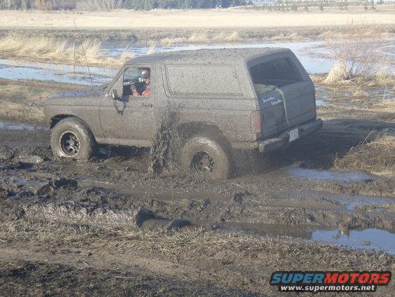 Mud-covered SUV stuck in mud, driver visible, rear tire deep in mud