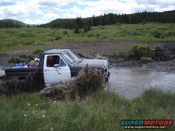 Two-tone pickup truck splashing through a muddy creek with people in the bed