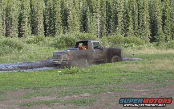 Mud-splattered pickup truck driving through a muddy puddle, with trees in the background
