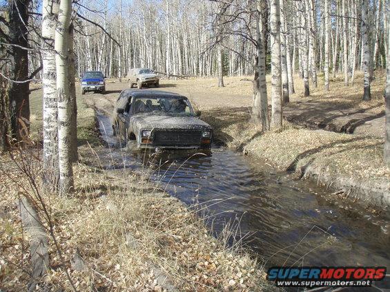 Muddy SUV driving through a stream with two other vehicles behind it on a dirt path in a forest